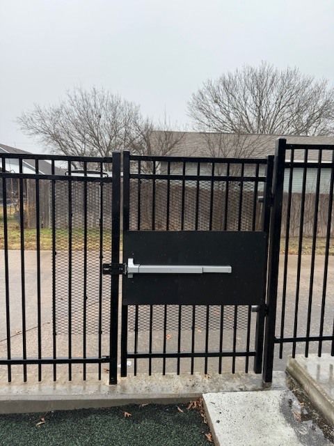 Black metal gate with a pedestrian door and silver push bar in a fenced outdoor area.