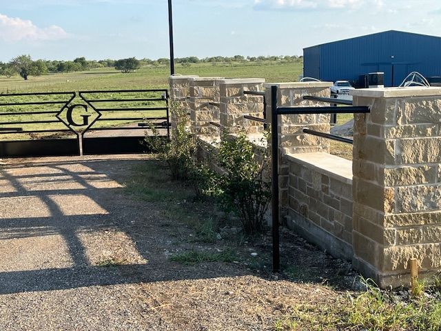 Stone gate and fence opening onto a gravel driveway with green fields beyond under a blue sky