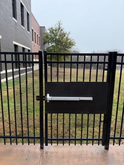 Black metal gate with a silver horizontal bar latch beside a brick building and grassy path.