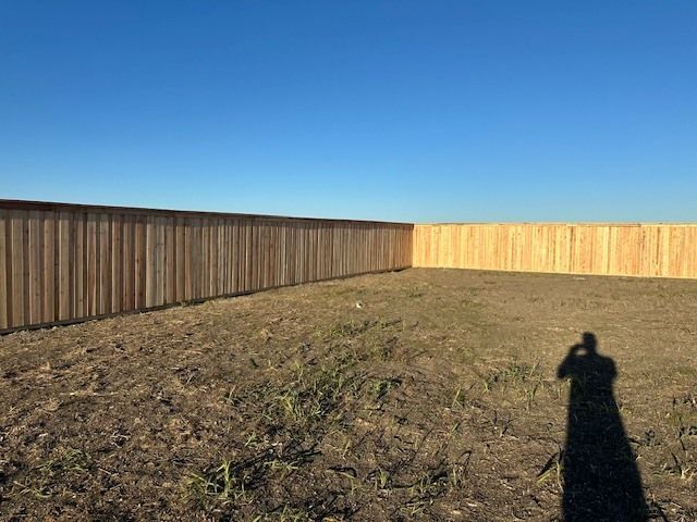 Empty dirt yard enclosed by wooden fences under a clear blue sky, with a long shadow in the foreground
