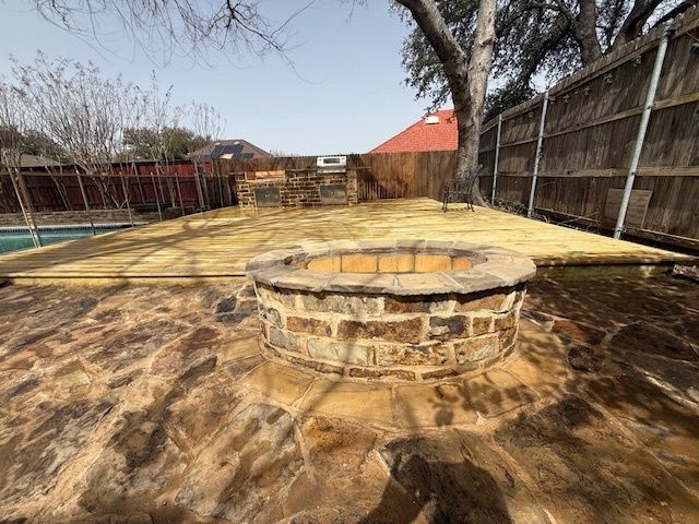 Backyard patio with a circular stone fire pit, wooden deck, and fenced yard under a clear sky