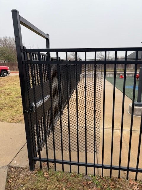 Black metal gate at a pool entrance beside a concrete walkway, with a handrail and water beyond.