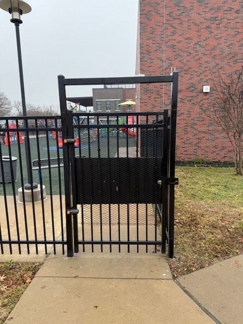Black metal gate with a walk-through pedestrian door beside a brick building and sidewalk.