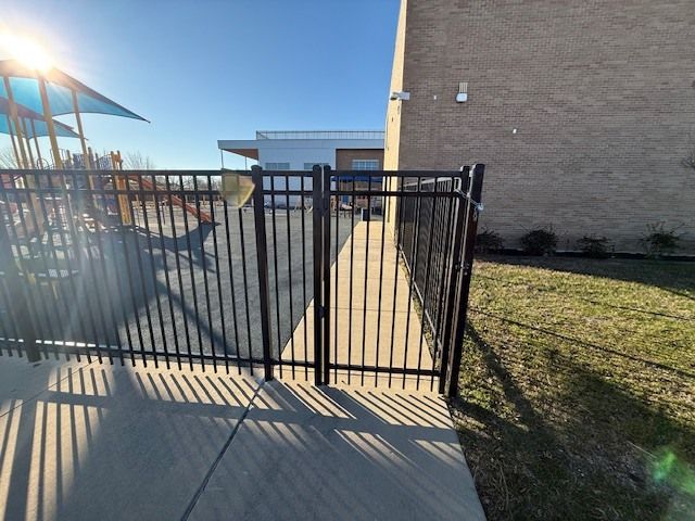 Black metal gate blocking a concrete walkway beside a brick building and grassy lawn in bright sunlight