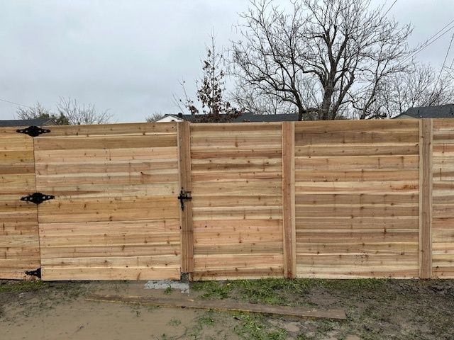 Wooden privacy fence with gates along a dirt lot under bare trees.