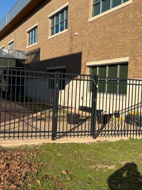 Brick building with black metal fence, ramp, and green double doors beside a grass lawn.
