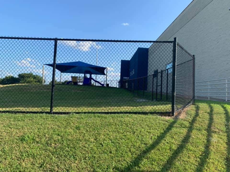 Chain-link fence and grassy hill beside a blue canopy tent and building under a clear sky.