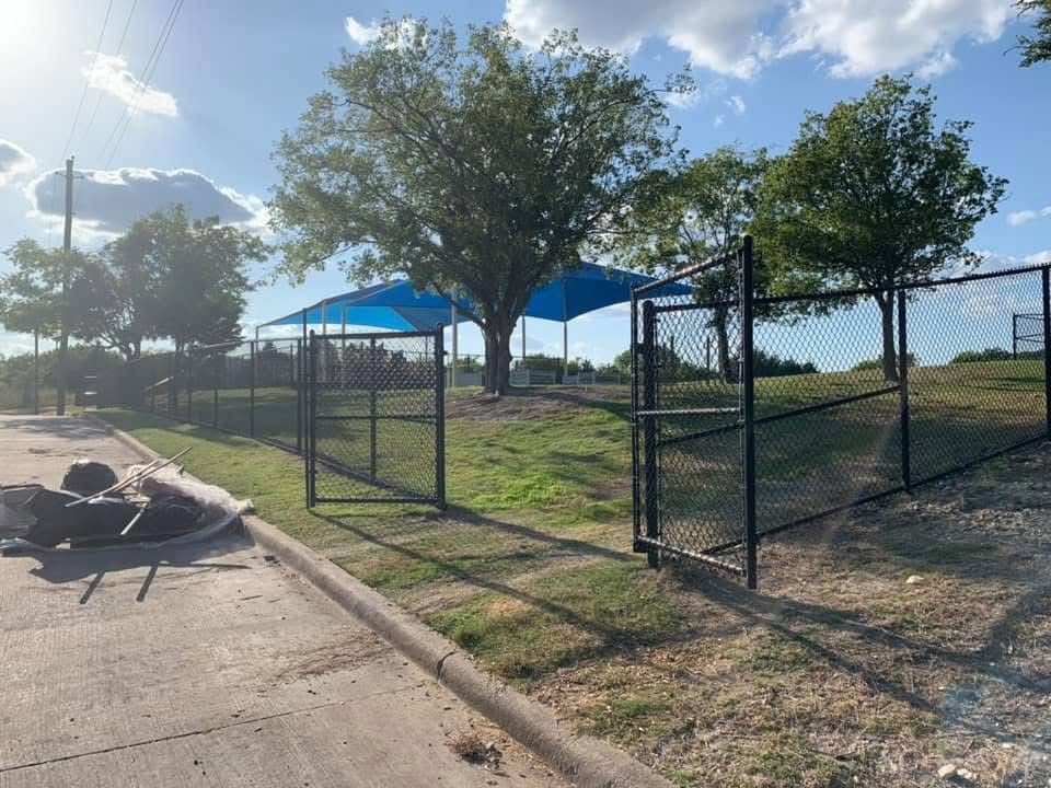 Chain-link dog park with blue shade canopies, grass, and a paved path on a sunny day