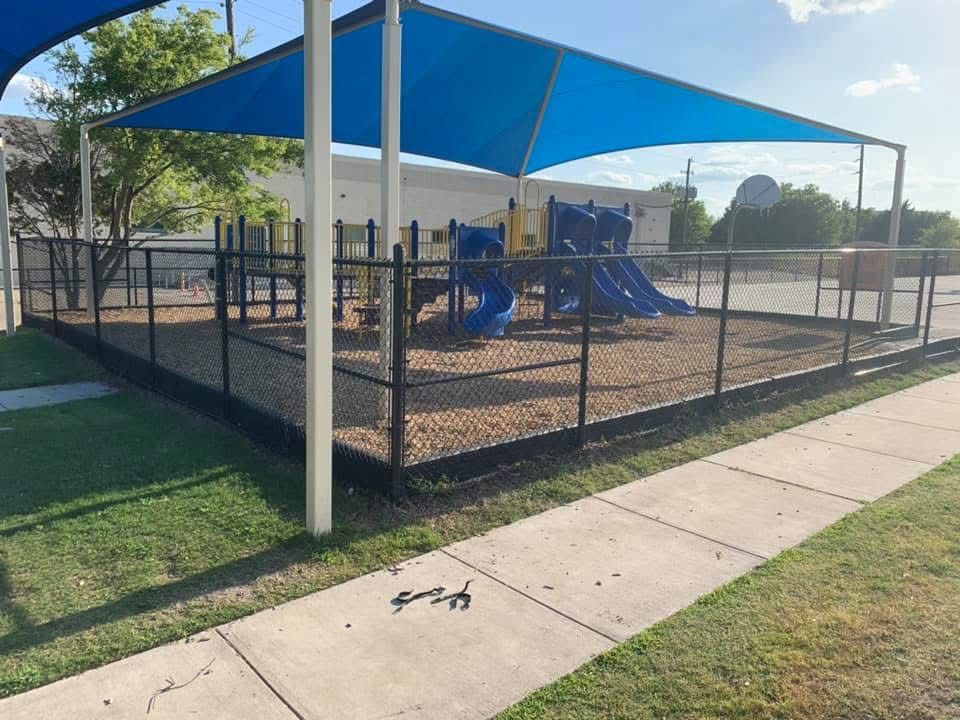 Outdoor playground with blue shade canopy, fenced play area, and blue slides beside a sidewalk and grass