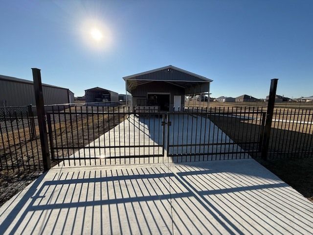 Gated concrete driveway leading to a house under a bright sun, with long fence shadows.
