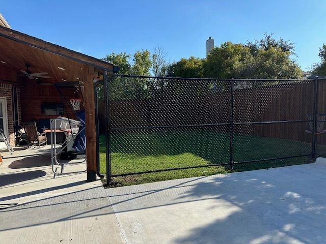 Backyard patio with black chain-link fence, green grass, and a shaded covered area with a grill