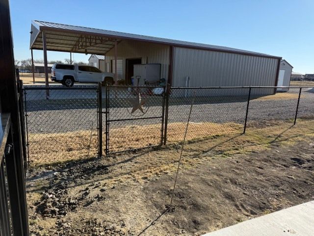 Rural yard with chain-link gate, metal carport, and white pickup truck on a dirt lot under a clear sky