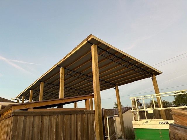 Wooden pergola-style outdoor structure with a sloped roof beside a fenced yard at sunset