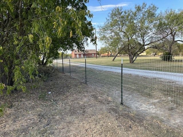 Wire fence along a gravel road beside trees and a rural house in the distance.