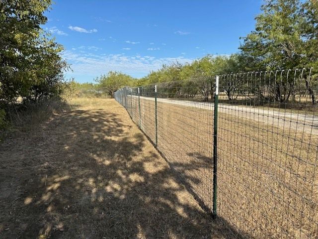 Chain-link fence beside a dirt road under a clear blue sky, with trees and dry grass.