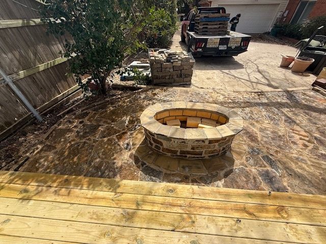 Backyard patio with circular stone fire pit, wooden deck, tree, and parked truck in the background