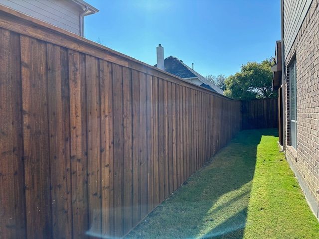 Narrow grassy side yard between a tall wooden fence and a brick house under a blue sky