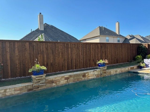 Backyard pool with clear blue water, wooden privacy fence, stone edging, and potted flowers under sunny sky