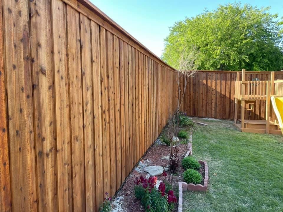Tall wooden backyard fence beside a narrow garden bed with flowers, lawn, and a patio deck in sunlight