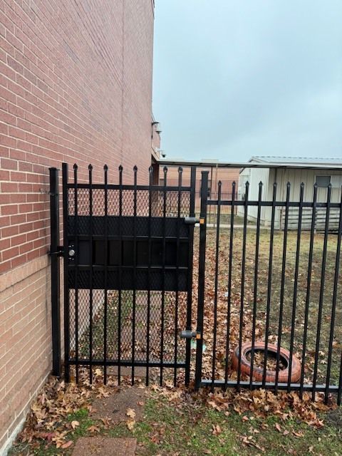 Black metal gate beside a red brick wall, with a mailbox and fallen leaves in a fenced yard.