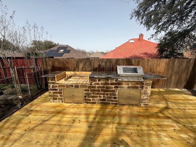 Backyard patio with a brick outdoor grill and wooden deck, fenced yard and houses in the background