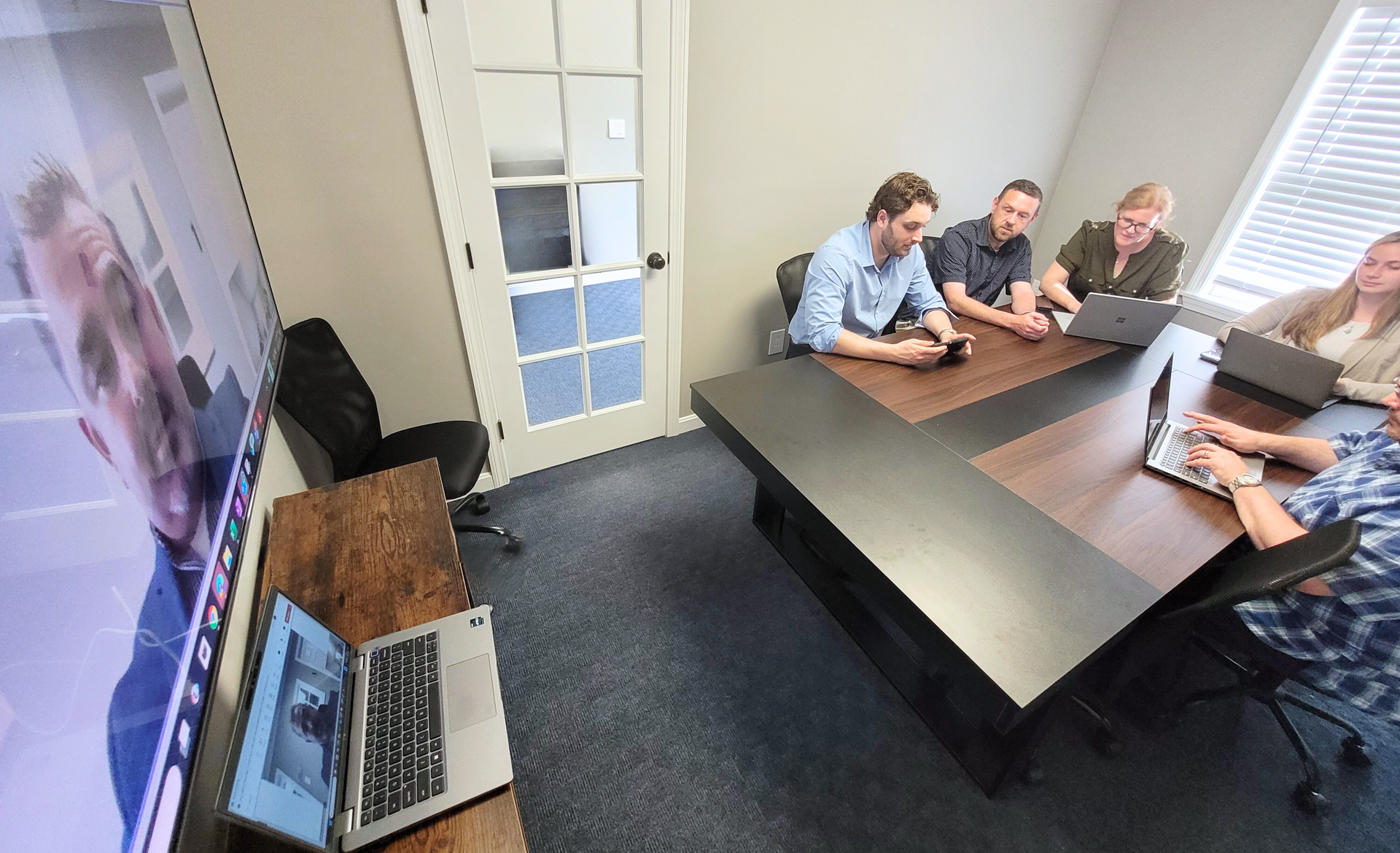 People in an office conference room sitting around a table with laptops while video calling someone on a large screen.
