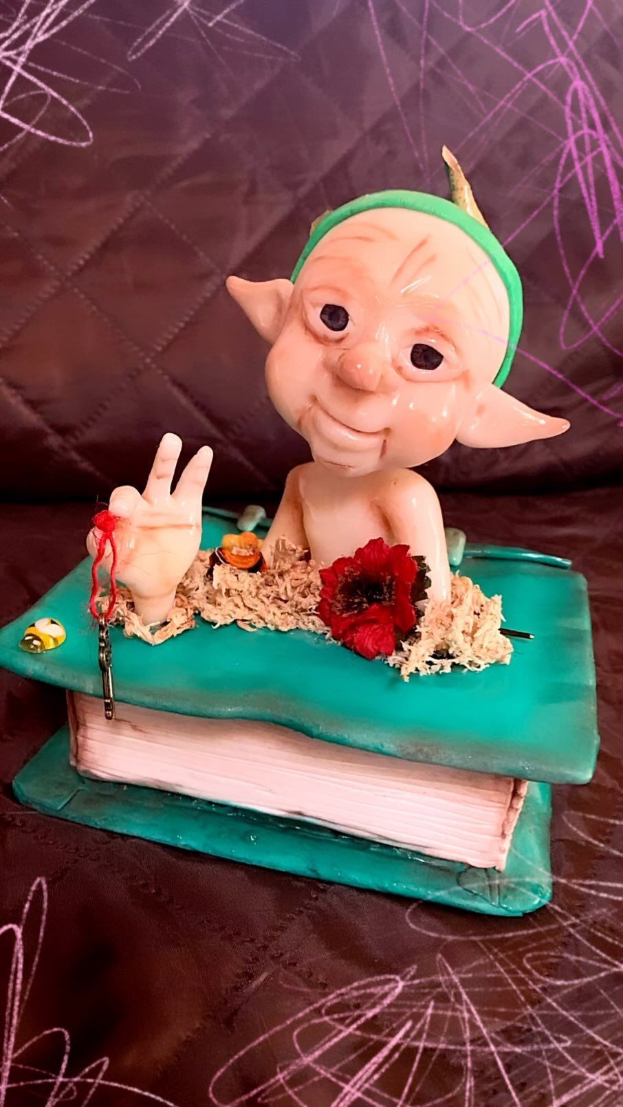 Pequeña figurita sobre un pastel color turquesa, rodeada de flores, sobre un fondo rosa oscuro.