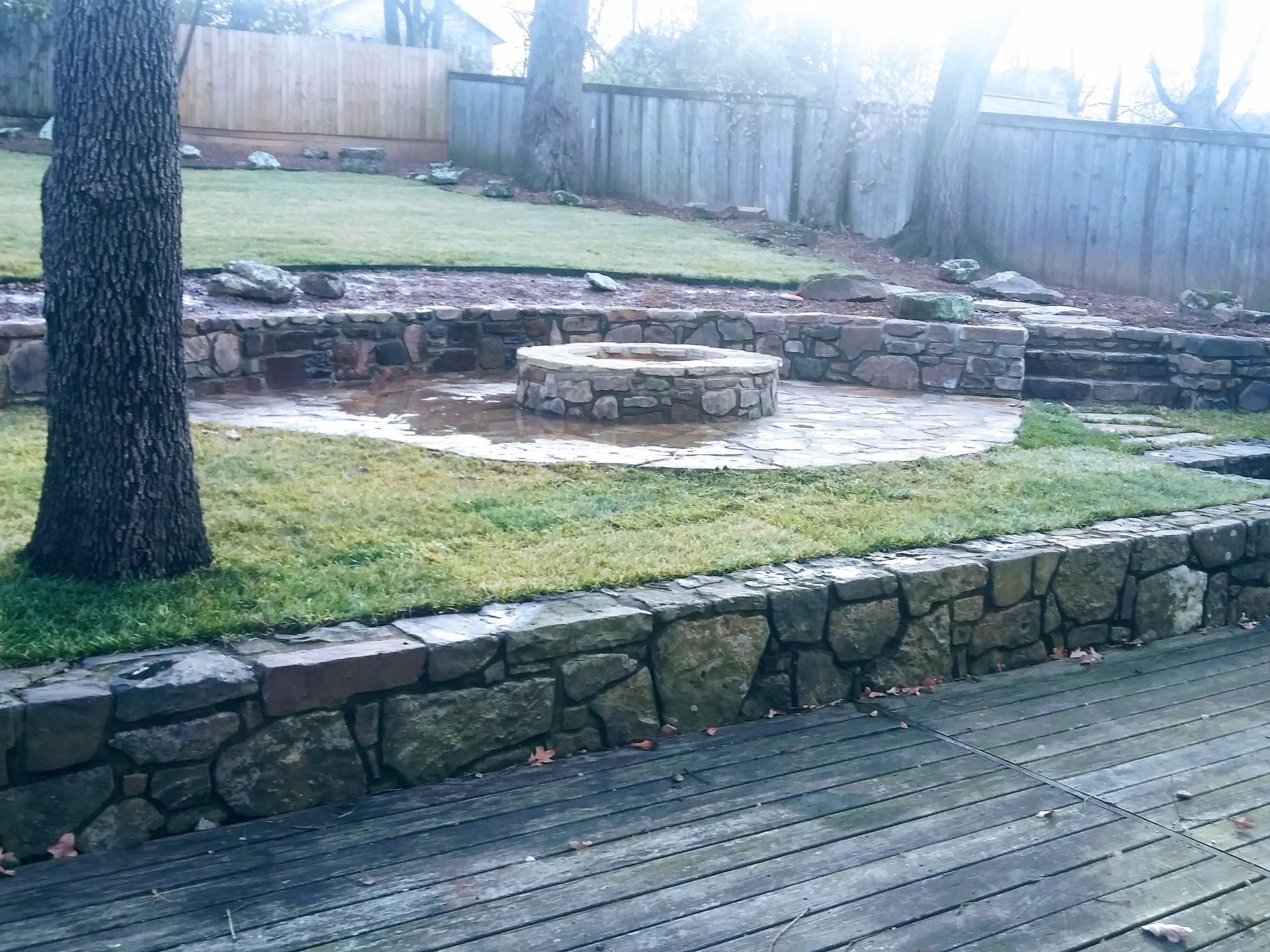 Stone fire pit area in a backyard, with stone retaining walls, wooden deck, and green grass.