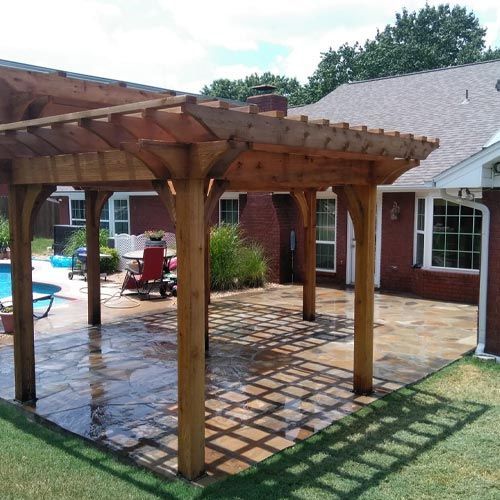 Wooden pergola over a stone patio with a pool in the background and a brick house.