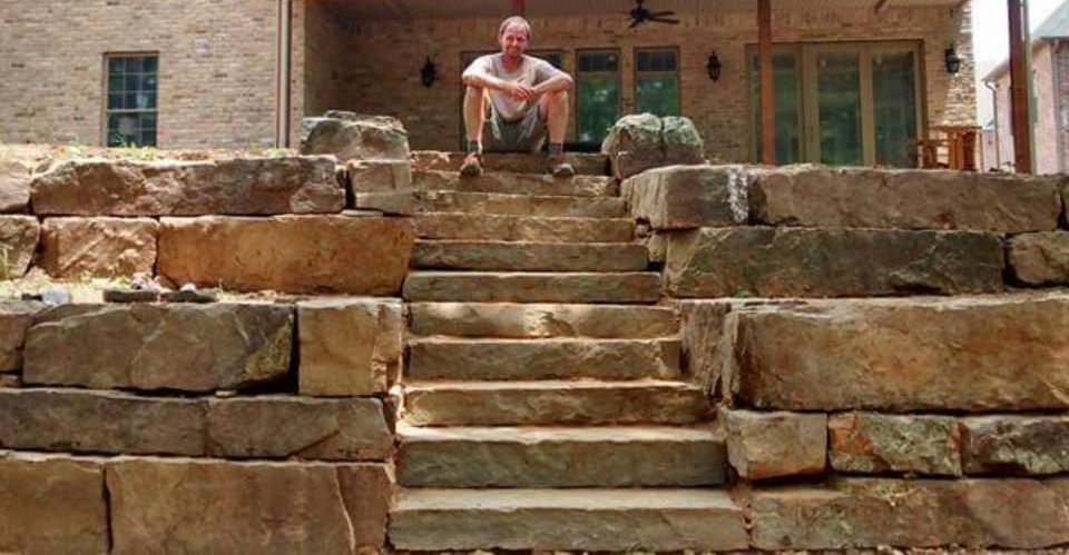 Man seated on stone steps, part of a tiered retaining wall. A building is visible in the background.