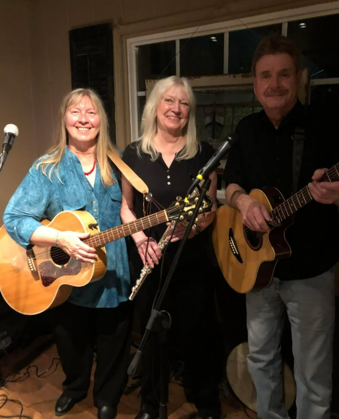 Three musicians stand indoors holding guitars and a flute, smiling at the camera.