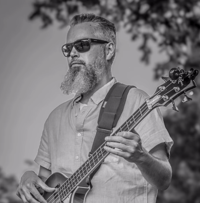 Man wearing sunglasses playing a bass guitar outdoors, in black and white.
