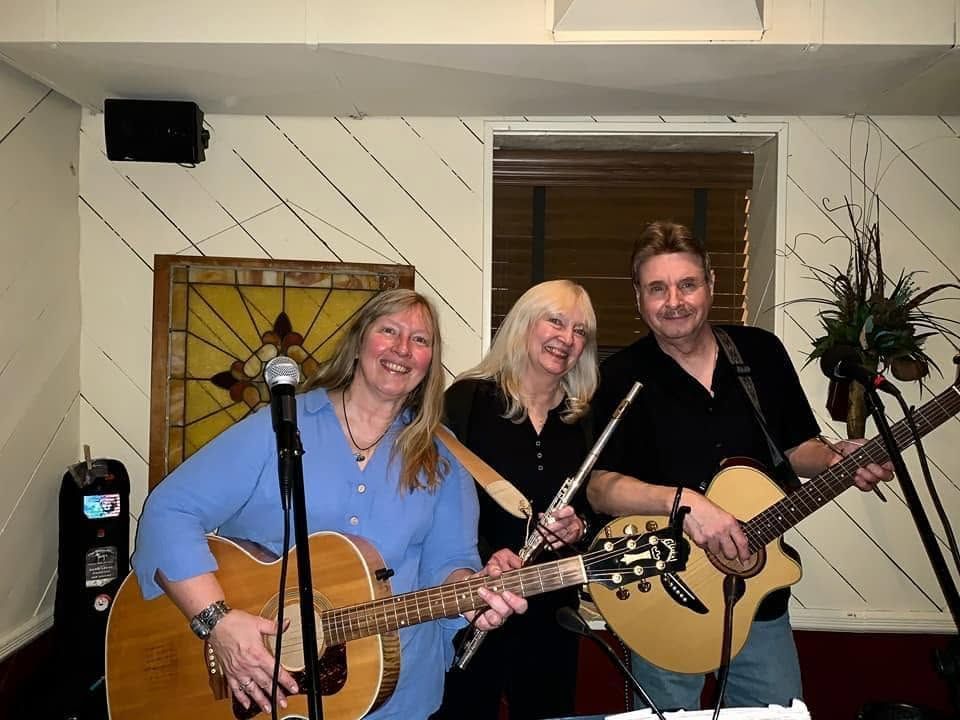 Three musicians smiling and playing acoustic guitars indoors before a striped wall