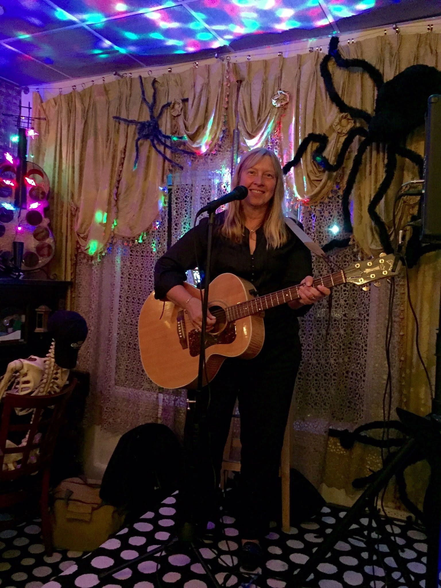 Musician playing acoustic guitar on a small stage with colorful lights and wall art behind her