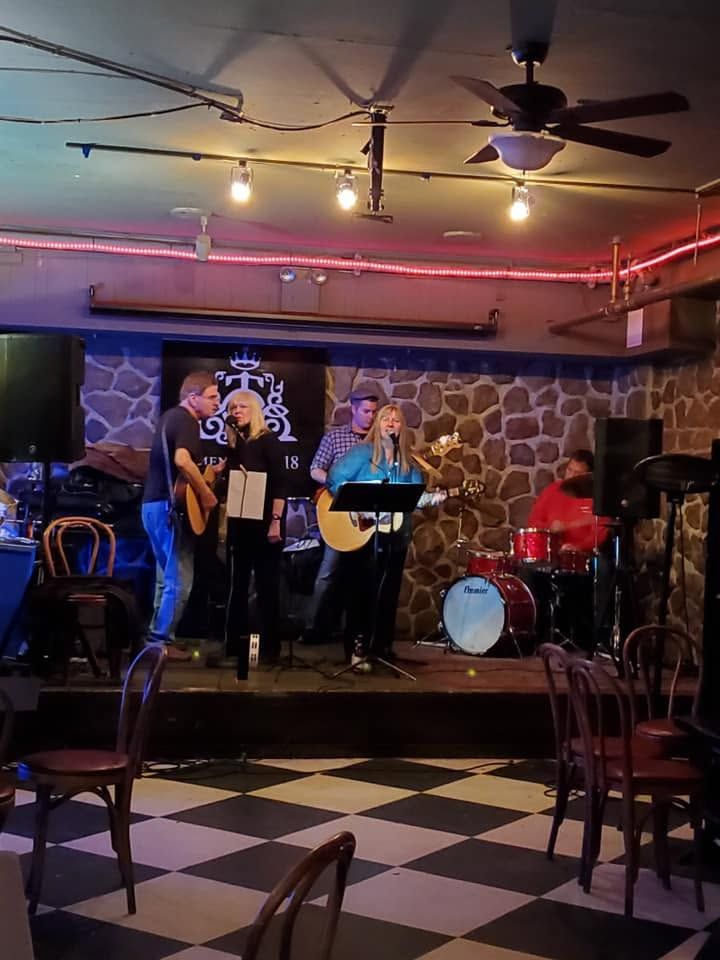Live band performing on a small stage in a dim bar with stone wall, pink neon lights, and checkered floor.