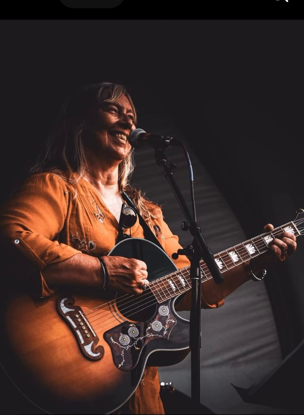 Singer playing guitar and singing into a microphone on a dark stage, wearing an orange jacket