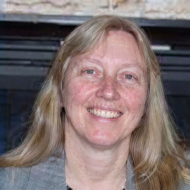 Smiling woman with long blonde hair in a gray top, seated indoors in front of a dark fireplace Smiling woman with long blonde hair in a gray top, seated indoors in front of a dark fireplace