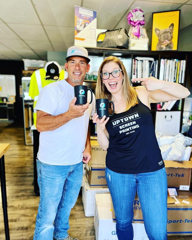 Two smiling people holding mugs in a storage room with boxes and shelves behind them