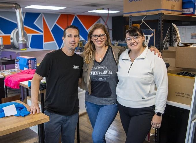 Three people posing in a cluttered office with boxes, tables, and colorful wall art.