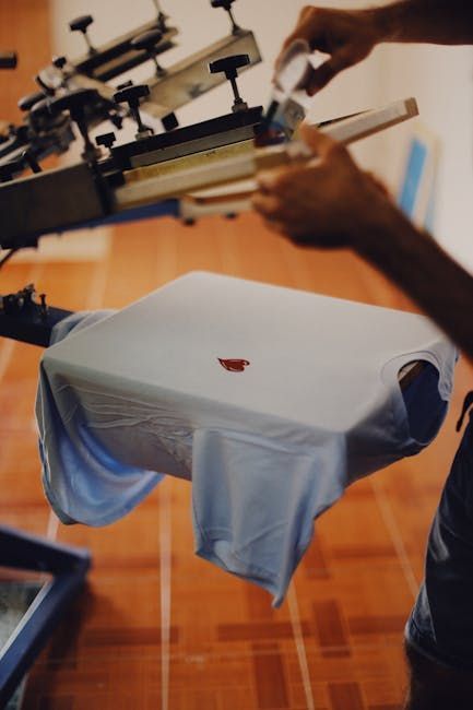 Person screen printing a red design on a light blue t-shirt with a printing machine.