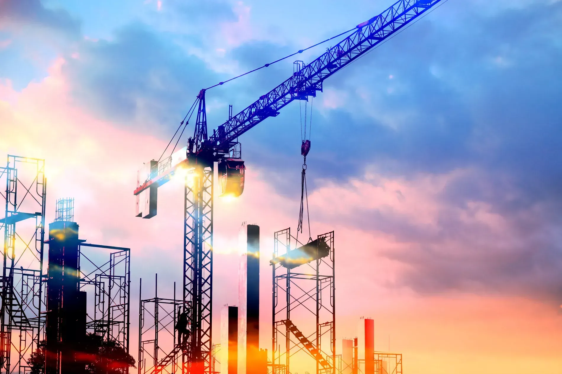 Construction site at sunset, crane lifting a beam; silhouettes against a colorful sky.