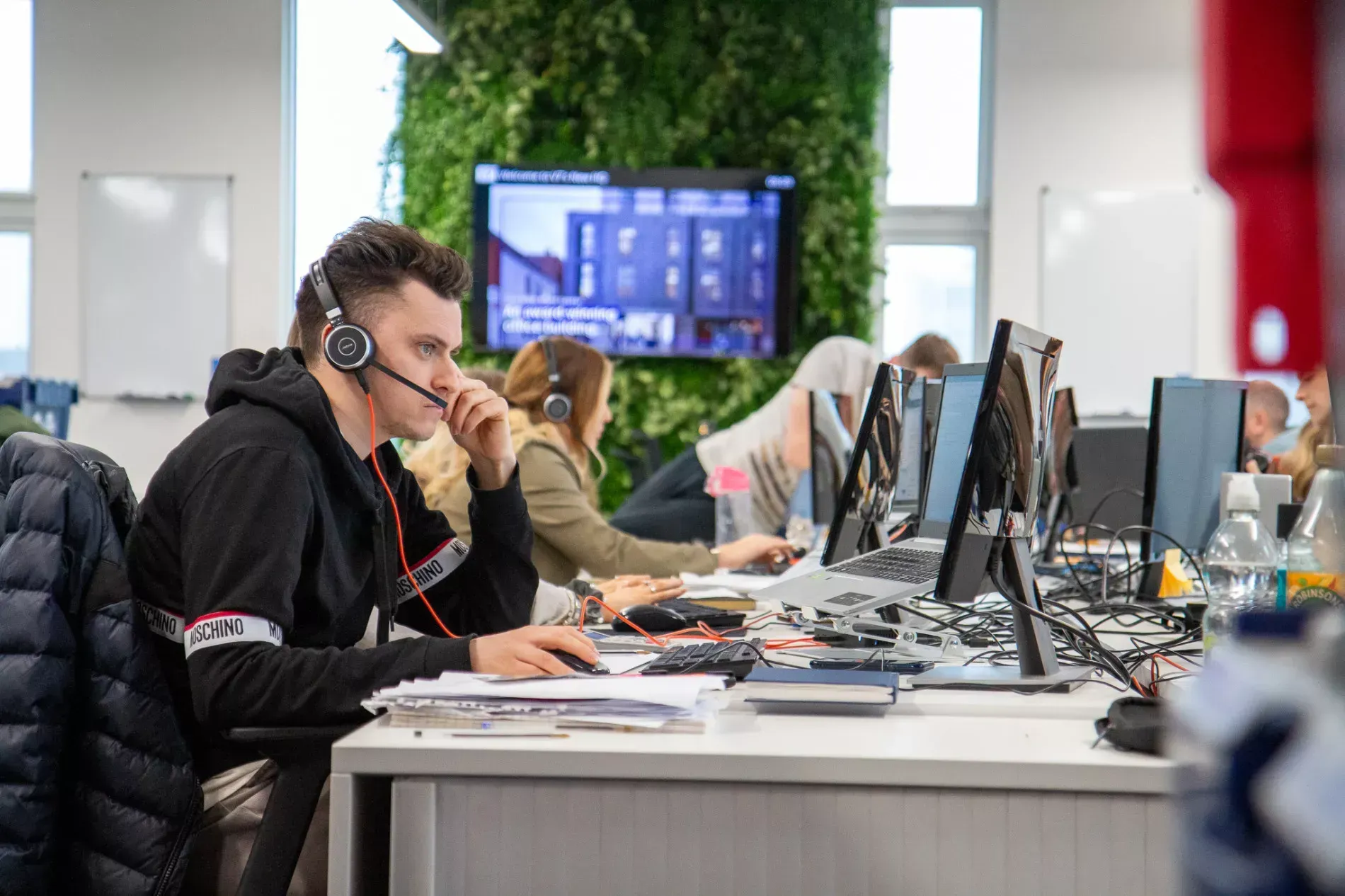 Office workers at desks, wearing headsets, with computers and green wall in the background.