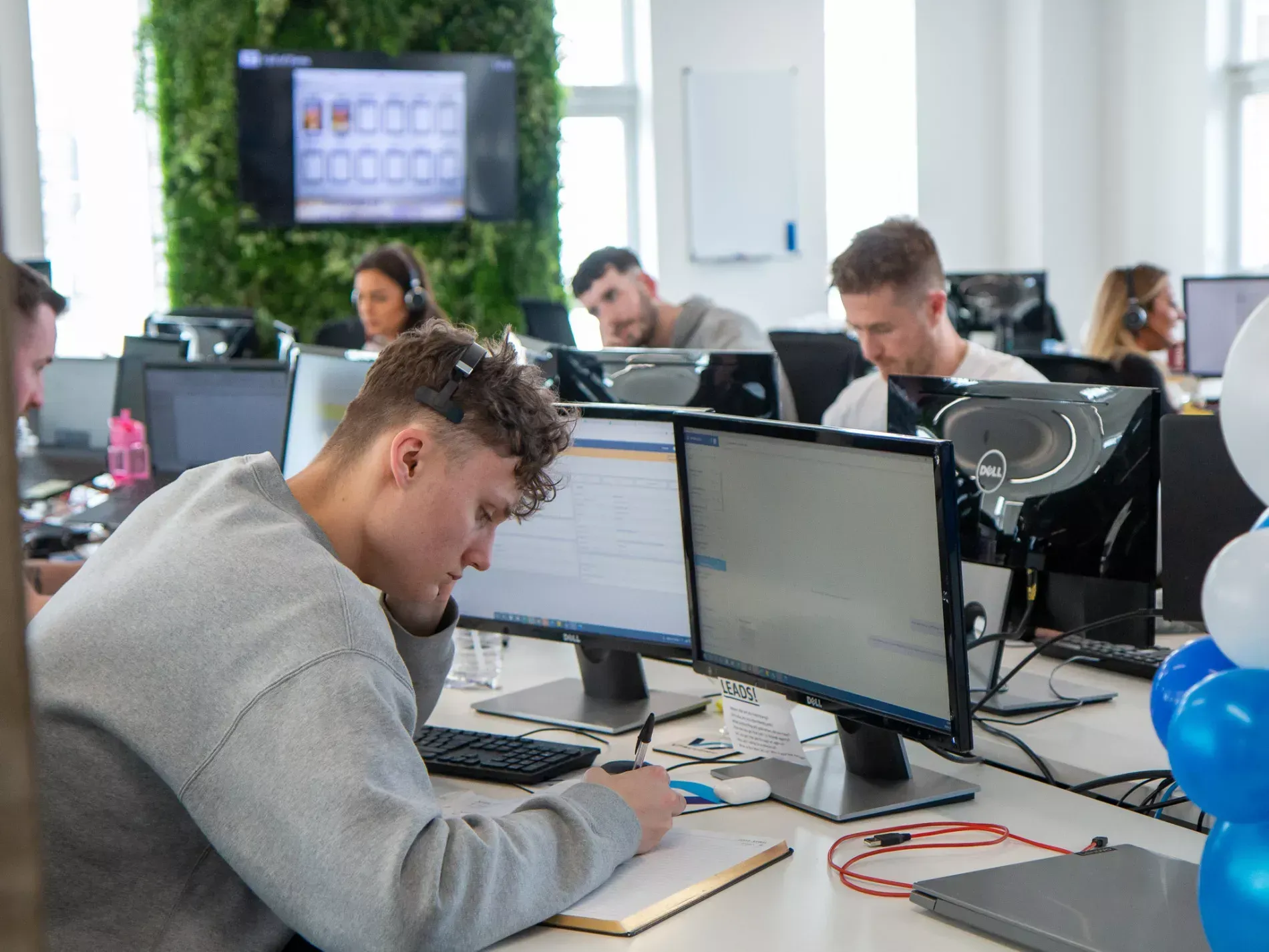 Office with several people working at computers; some wearing headsets, focused.