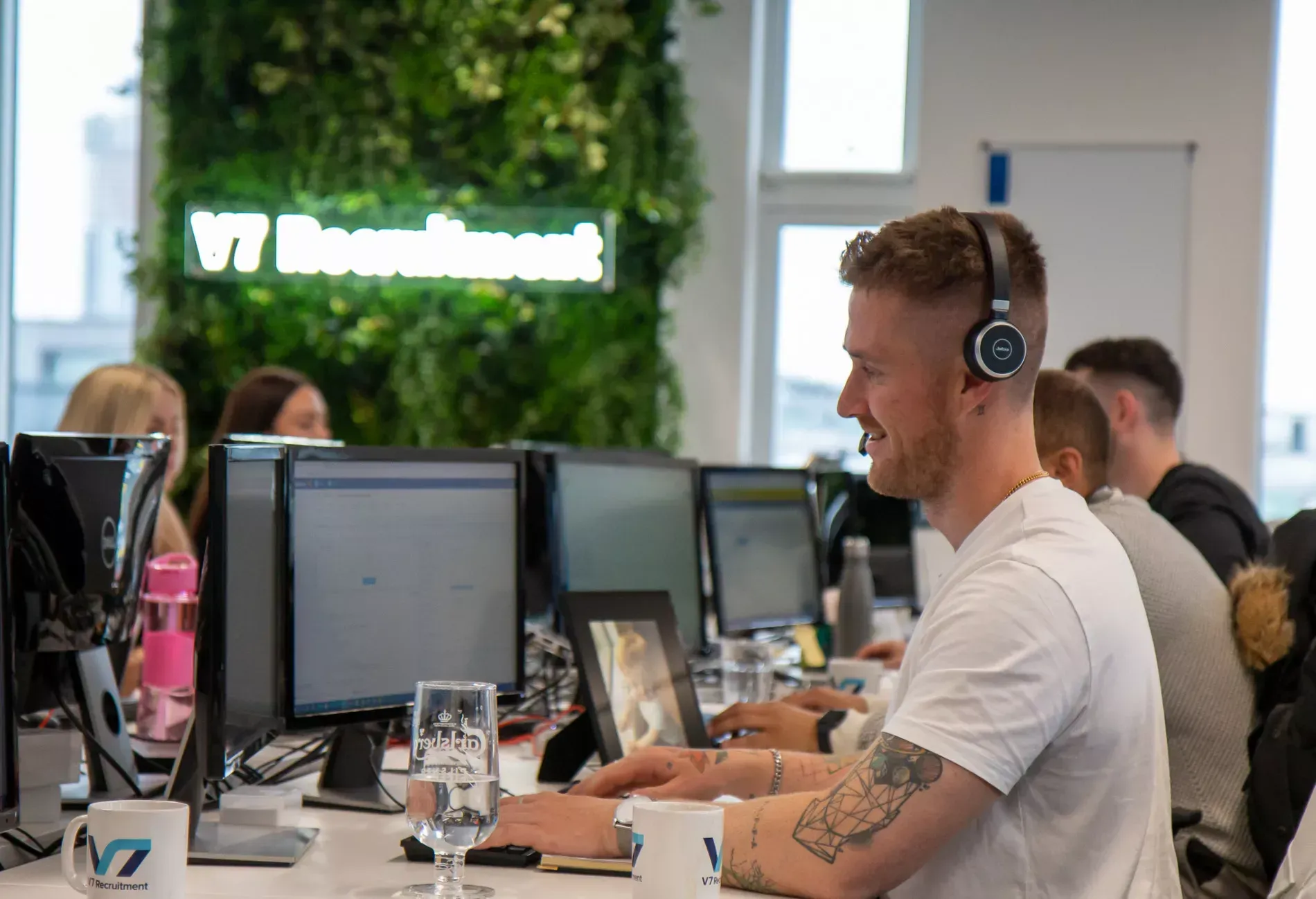 A man with headphones smiles while working at a computer in an office. Others work nearby.