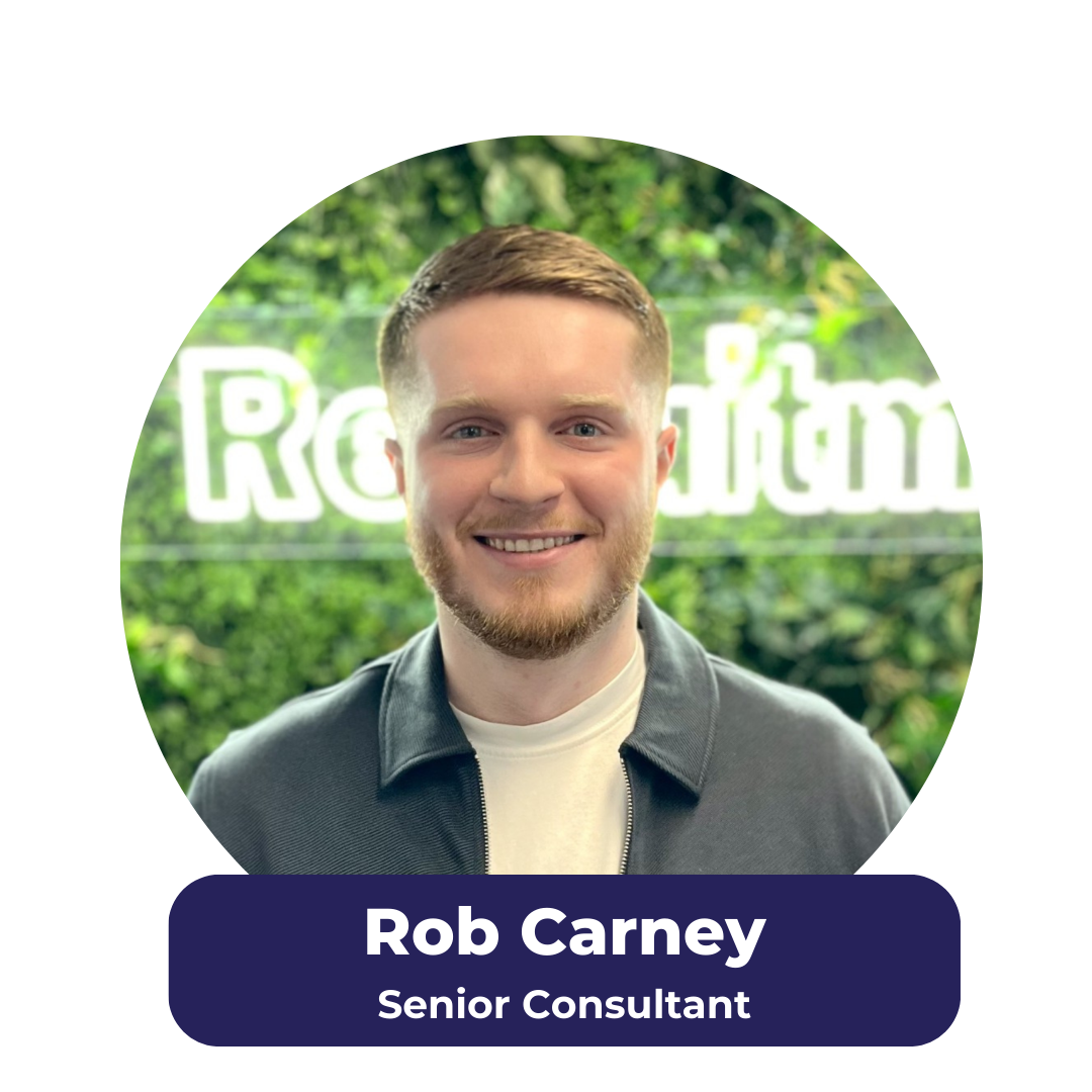 Rob Carney, a senior consultant, smiles in front of a neon sign and greenery.