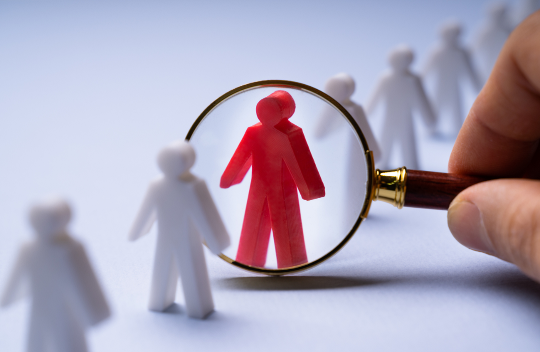 Row of white people figures, one person in the middle is red with a magnifying glass over him