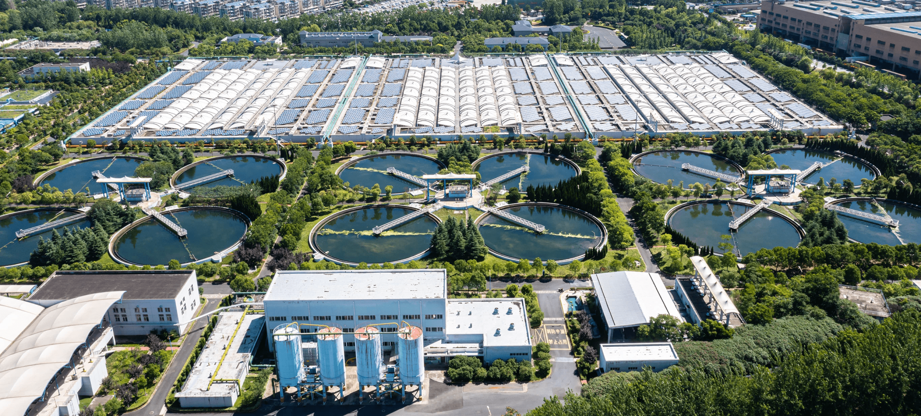 Aerial view of a water treatment facility with large basins and rectangular structures surrounded by trees and buildings.