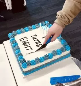 Person cutting a square cake with blue and gray frosting, which says