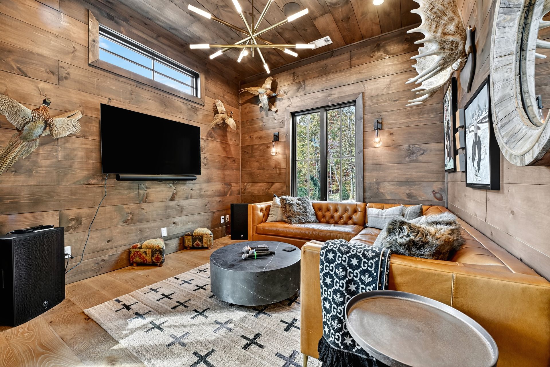 Cozy living room with wood-paneled walls, leather sectional, rug, large TV, antlers, and a modern chandelier.