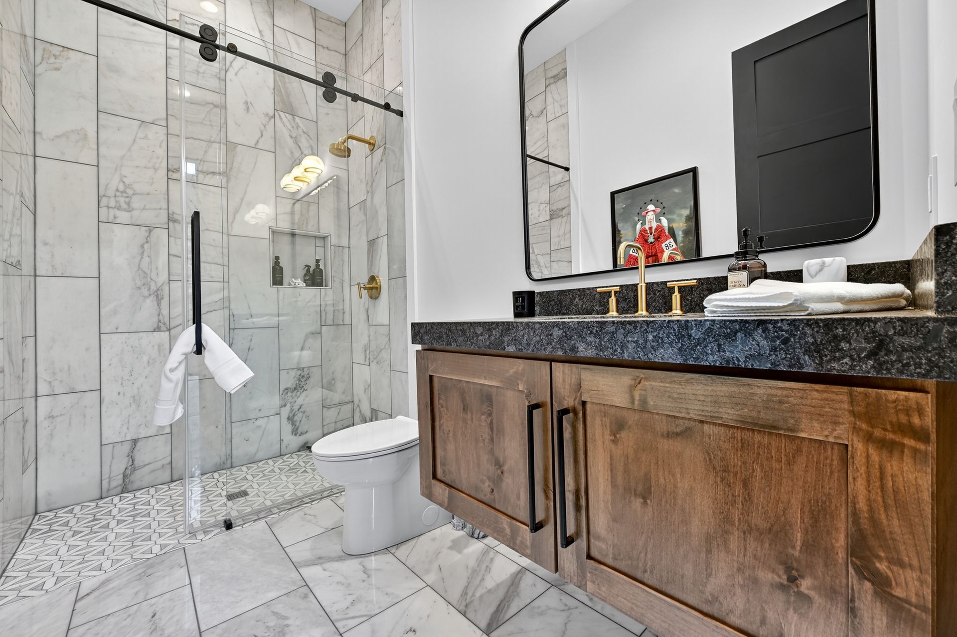 Bathroom with marble tiles, glass shower, wooden vanity, black countertop, gold fixtures.